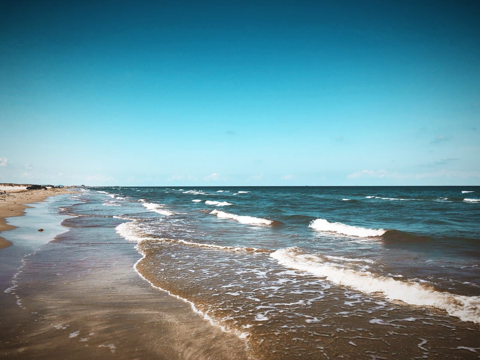 Sunny day at Mustang Island State Park — turquoise Gulf waves rolling onto packed Texas sand, deep blue sky and clean horizon, looking south down the beach.