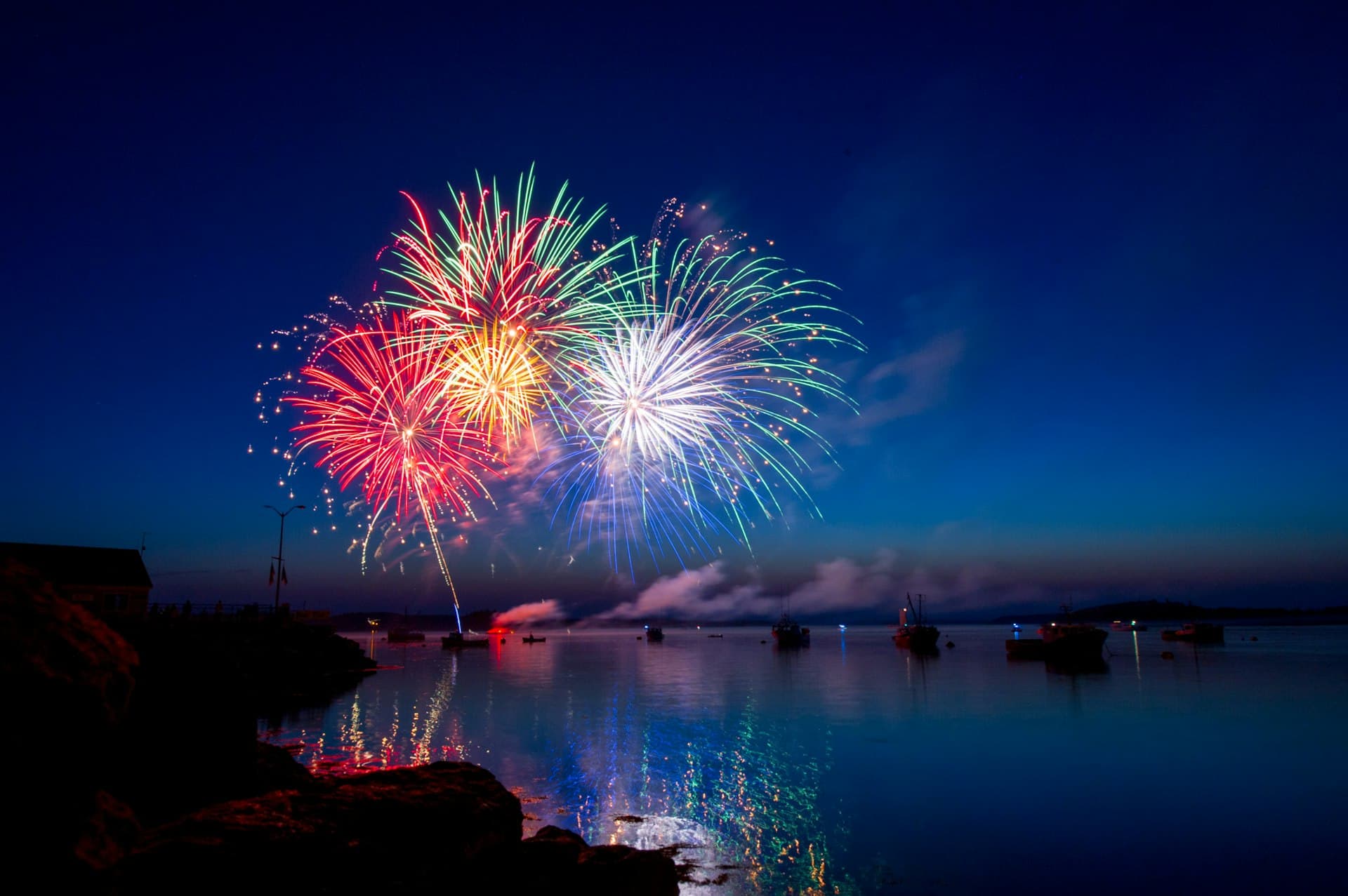 Red, white, and blue fireworks bursting over a calm harbor with fishing boats moored on the water at dusk