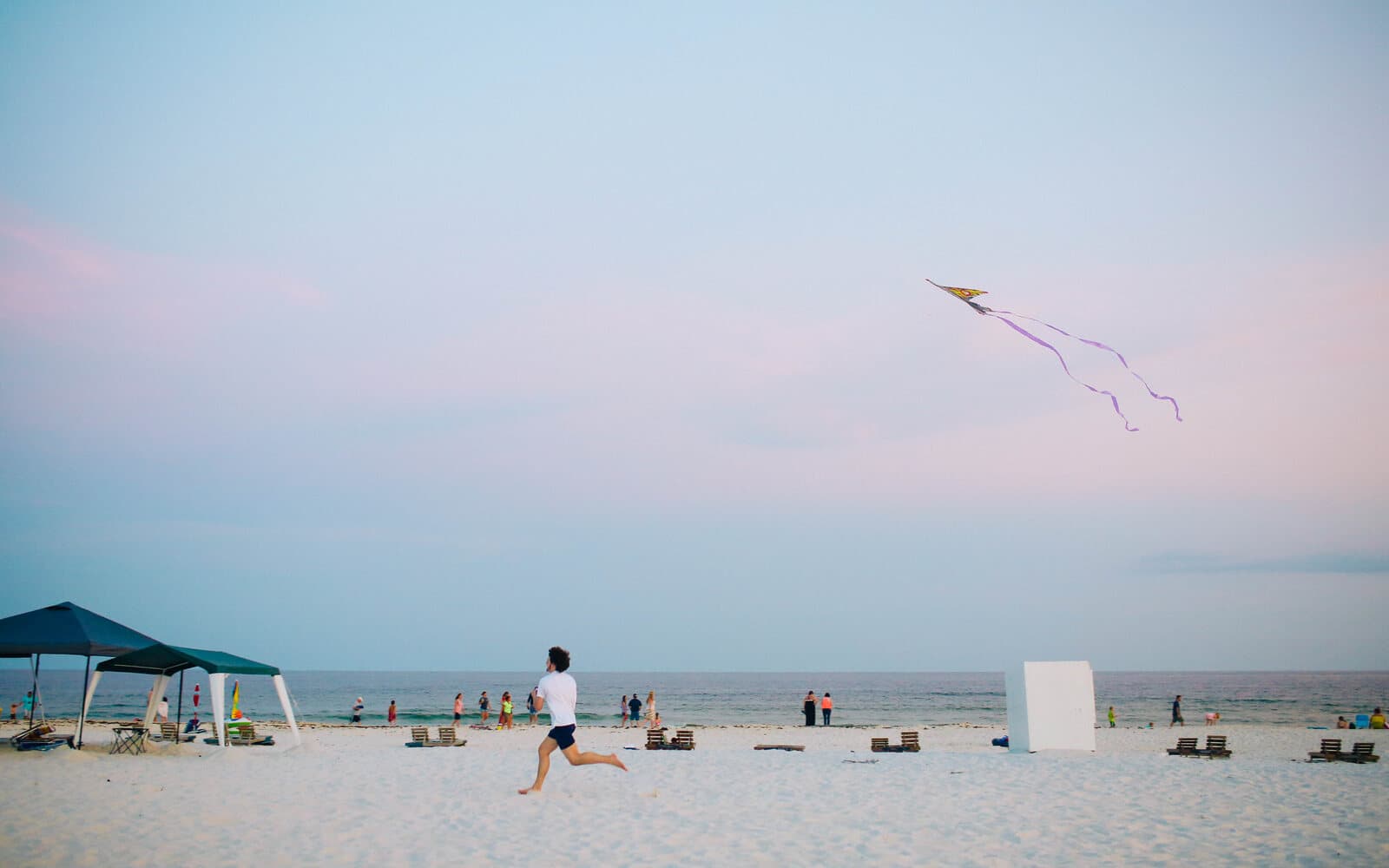 Open sandy beach with kites flying in a pastel sky above a runner and beach umbrellas.