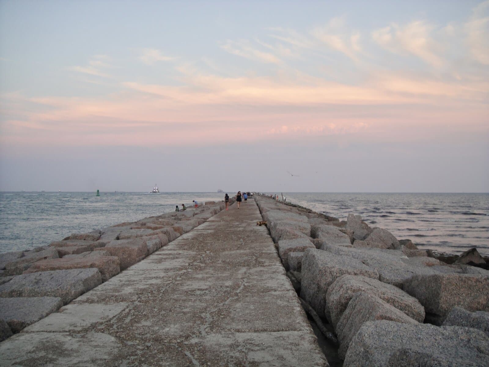 The Port Aransas South Jetty granite walkway at sunset — anglers and walkers along the rocks, a sailing ship passing in the distance through the channel under a pastel sky.