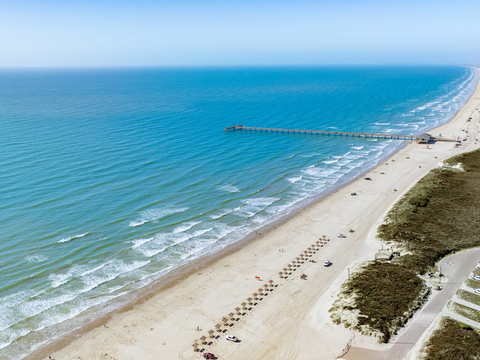Aerial view of Port Aransas Beach — drive-on Gulf shore lined with Beach Bub umbrella rentals, the fishing pier reaching offshore, dunes and a quiet beach access road at right.