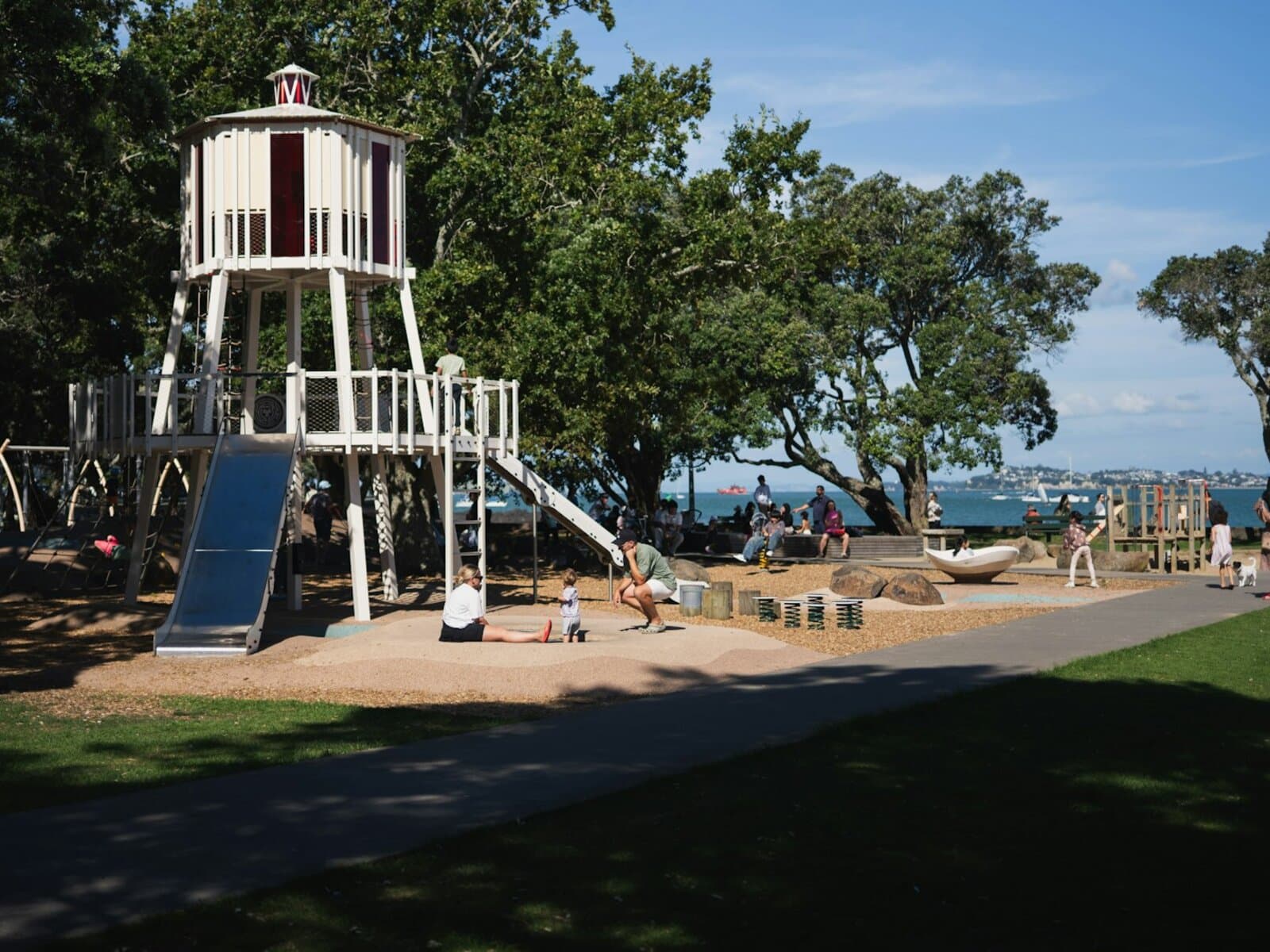 Coastal park playground in Port Aransas: a tall white wooden tower with red trim styled like a lighthouse, with a metal slide and climbing structures, oak trees around it and the marina water visible beyond.