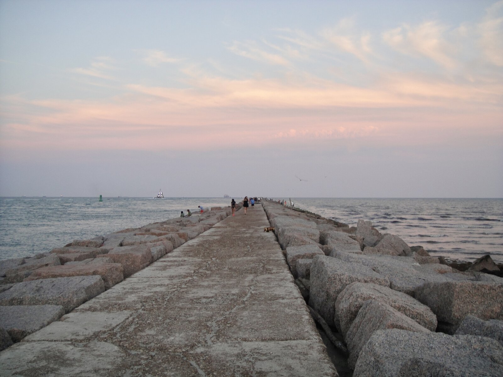 The Port Aransas South Jetty granite walkway at sunset — anglers and walkers along the rocks, a sailing ship passing in the distance through the channel under a pastel sky.